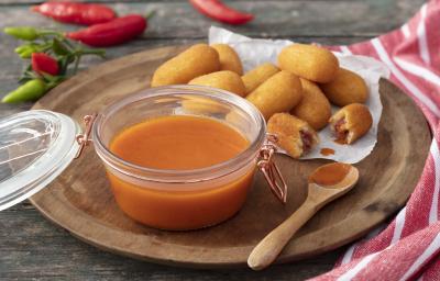 Fotografia em tons de vermelho em uma bancada de madeira escura, um pano vermelho com listras brancas, uma tábua de madeira escura com um pote com tampa hermética de vidro e o molho de pimenta dentro dele. Ao lado, bolinhos de carne seca para acompanhar. Fotografia em tons de vermelho em uma bancada de madeira escura, um pano vermelho com listras brancas, uma tábua de madeira escura com um pote com tampa hermética de vidro e o molho de pimenta dentro dele. Ao lado, bolinhos de carne seca para acompanhar.