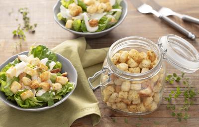 Fotografia em tons de verde em uma bancada de madeira clara, um pano verde, dois bowl com os croutons com a salada de alface e molho de iogurte. Ao centro, um pote de vidro hermético com vários croutons dentro dele. Fotografia em tons de verde em uma bancada de madeira clara, um pano verde, dois bowl com os croutons com a salada de alface e molho de iogurte. Ao centro, um pote de vidro hermético com vários croutons dentro dele.