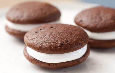 Fotografia em tons de preto em uma bancada de madeira clara, um prato redondo branco raso com três sanduicinhos de chocolate recheados com marshmallow. Fotografia em tons de preto em uma bancada de madeira clara, um prato redondo branco raso com três sanduicinhos de chocolate recheados com marshmallow.