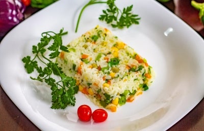 Em um prato de porcelana branco, há um porção colorida de arroz amarelo, milho verde, ervilha, decorados com tomate cereja e galhos de coentro. Em um prato de porcelana branco, há um porção colorida de arroz amarelo, milho verde, ervilha, decorados com tomate cereja e galhos de coentro.