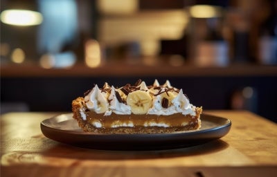 Em cima de uma mesa, prato com fatia de banoffee com massa crocante, doce de leite com chantilly, decorado com chantilly, bananas em rodelas e raspas de chocolate. Em cima de uma mesa, prato com fatia de banoffee com massa crocante, doce de leite com chantilly, decorado com chantilly, bananas em rodelas e raspas de chocolate.