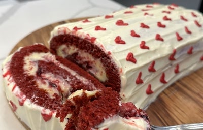 Fotografia em tons claros com um bolo de rolo de frutas vermelhas ao centro. A massa do bolo tem cor vermelha, com recheio branco e cobertura branca com decorações de coração.