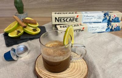Fotografia em tons de amarelo e azul com uma xícara de vidro ao centro. Dentro da xícara, que está enfeitada com uma fatia de laranja, existe uma mistura de café com suco de laranja. Ao fundo existe fatias de laranja e duas caixas de cápsulas NESCAFÉ Fotografia em tons de amarelo e azul com uma xícara de vidro ao centro. Dentro da xícara, que está enfeitada com uma fatia de laranja, existe uma mistura de café com suco de laranja. Ao fundo existe fatias de laranja e duas caixas de cápsulas NESCAFÉ