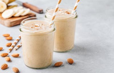 Fotografia em tons claros de uma mesa de cimento queimado claro. Sobre a mesa tem dois copos de vidro com milkshake de coco e amêndoas, com lascas de amêndoas sobre cada um. Dentro de cada copo tem um canudo branco e marrom, ao lado de amêndoas.