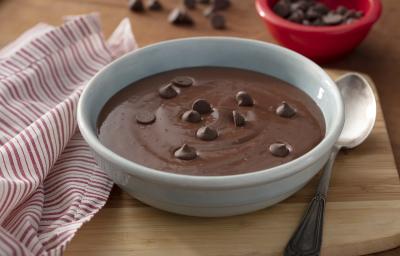 Fotografia em tons de vermelho em uma bancada de madeira clara, um pano branco com listras vermelhas, uma tábua de madeira com um recipiente redondo fundo com a sobremesa mingau de chocolate dentro dele e algumas gotas de chocolate para decorar.