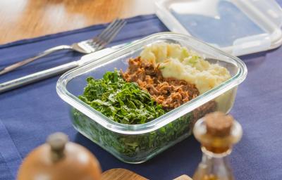 A fotografia mostra uma marmita em pote de vidro com carne desfiada ao meio, purê de batata-doce cremoso no canto superior direito e couve refogada verde no canto esquerdo.