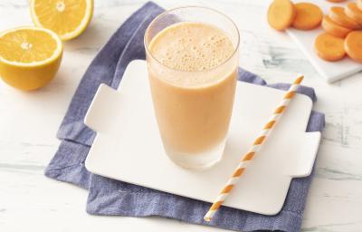 Fotografia em tons de laranja em uma bancada de madeira clara com um pano azul e uma tábua branca ao centro com um copo de vidro com a bebida refrescante de laranja e cenoura. Ao lado, um canudo listrado laranja, algumas fatias de cenoura e uma laranja.