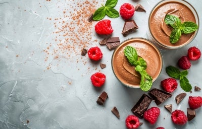 Fotografia vista de cima de dois potes de vidro sobre uma mesa de cimento queimado. Dentro do copos tem a sobremesa, finalizada com ganache e folhas de hortelã. Ao redor dos copos tem hortelãs, pedaços de chocolate e framboesas espalhadas. Fotografia vista de cima de dois potes de vidro sobre uma mesa de cimento queimado. Dentro do copos tem a sobremesa, finalizada com ganache e folhas de hortelã. Ao redor dos copos tem hortelãs, pedaços de chocolate e framboesas espalhadas.