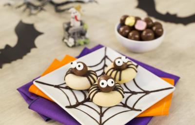 Imagem aproximada de um prato quadrado branco com três biscoitos em formatos de aranhas. A bancada está decora com papéis em tons roxo e laranja e alguns morcegos de mentira. Imagem aproximada de um prato quadrado branco com três biscoitos em formatos de aranhas. A bancada está decora com papéis em tons roxo e laranja e alguns morcegos de mentira.
