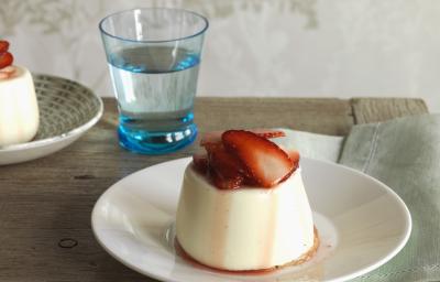 Fotografia de um flan de canela com calda de morangos em um prato branco fundo sobre uma mesa de madeira. Ao fundo, outro flan de canela e, ao lado, um copo d'água. Fotografia de um flan de canela com calda de morangos em um prato branco fundo sobre uma mesa de madeira. Ao fundo, outro flan de canela e, ao lado, um copo d'água.