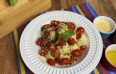 Fotografia em tons de amarelo e vermelho em uma bancada de madeira com um pano colorido em azul, vermelho e amarelo e um prato branco fundo em cima dele com o gnudi e molho de tomate por cima dentro do prato. Ao lado, potinhos com queijo ralado e azeite.