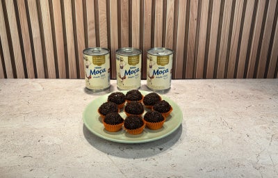 Prato verde-claro com brigadeiros cobertos com granulado de chocolate e latas de Leite Moça ao fundo.