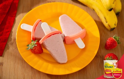 Picolés de morango e banana em forminhas com palito, sobre prato amarelo, com morangos e bananas ao redo.