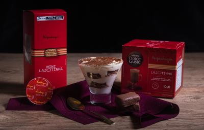 Fotografia em tons escuros de um recipiente de vidro com tiramisú sobre um pano vinho com uma colher de sopa e duas lajotinhas ao lado de uma embalagem de mini lajotinha e uma outra de dolce gusto lajotinha. Fotografia em tons escuros de um recipiente de vidro com tiramisú sobre um pano vinho com uma colher de sopa e duas lajotinhas ao lado de uma embalagem de mini lajotinha e uma outra de dolce gusto lajotinha.