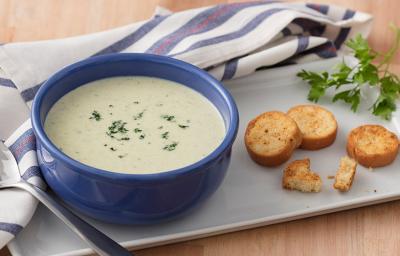 Fotografia em tons de azul e verde, com cumbuca azul ao centro com creme de legumes e salsa picada com torradinhas sobre travessa branca, ramo de salsa, brócolis e couve-flor decorando, guardanapo branco e azul com colher, sobre bancada de cor palha. Fotografia em tons de azul e verde, com cumbuca azul ao centro com creme de legumes e salsa picada com torradinhas sobre travessa branca, ramo de salsa, brócolis e couve-flor decorando, guardanapo branco e azul com colher, sobre bancada de cor palha.