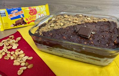 Fotografia em tons de amarelo e vermelho com uma travessa de sobremesa ao centro. Na travessa existe uma sobremesa a base de chocolate e coberta com pedações de amendoim e tabletes de chocolate ao leite. Ao fundo existe uma barra de chocolate fechada. Fotografia em tons de amarelo e vermelho com uma travessa de sobremesa ao centro. Na travessa existe uma sobremesa a base de chocolate e coberta com pedações de amendoim e tabletes de chocolate ao leite. Ao fundo existe uma barra de chocolate fechada.