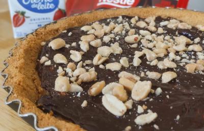 Fotografia de uma torta inteira charge ainda dentro da forma de alumínio. Por cima da torta tem amendoim, no fundo tem creme de leite e chocolate. A forma está sobre uma tábua de madeira.