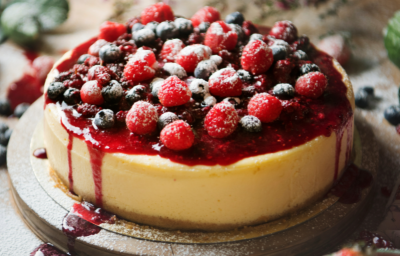 Imagem de um cheesecake coberto com uma calda vermelha e frutas vermelhas. Está disposto sobre uma tábua de madeira redonda, em cima de uma mesa de mandeira.