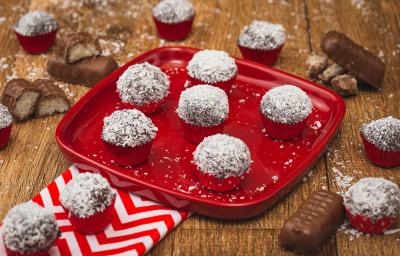 Imagem de uma prato vermelho com algumas unidades de Brigadeiro marrom passado em flocos de coco. A bancada está decorada com pedaços de bombons, um tecido vermelho e branco e mais brigadeiros. Imagem de uma prato vermelho com algumas unidades de Brigadeiro marrom passado em flocos de coco. A bancada está decorada com pedaços de bombons, um tecido vermelho e branco e mais brigadeiros.