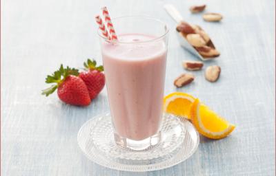 Fotografia em tons de rosa em uma bancada de madeira cinza com uma taça de vidro com a vitamina de morango com laranja dentro dele. Ao fundo, morangos, laranja e castanhas espalhados pela mesa. Fotografia em tons de rosa em uma bancada de madeira cinza com uma taça de vidro com a vitamina de morango com laranja dentro dele. Ao fundo, morangos, laranja e castanhas espalhados pela mesa.