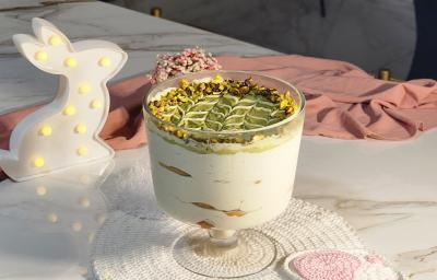 Fotografia de uma taça de vidro servido com camadas de creme, biscoito e pasta de pistache. Ao fundo, um pano rosa e uma luminária em formato de coelho. Fotografia de uma taça de vidro servido com camadas de creme, biscoito e pasta de pistache. Ao fundo, um pano rosa e uma luminária em formato de coelho.