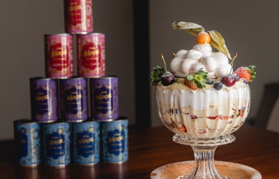 Foto da receita de pavlova na taça, montada em uma taça de cristal com camadas de merengue, creme e frutas amarelas. Servida ao lado de uma pilha de Latas de Leite moça