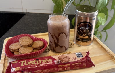 Foto da receita de Frappuccino de Chocolate com Biscoito Tostines, servido em um copo com canudo, sobre uma bandeja de madeira com Biscoitos Tostines Cappuccino soltos e o pacote à frente e, do lado direito, uma lata de Nescafé Gold