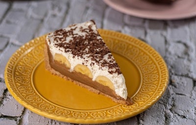 Em cima de uma mesa, prato com fatia de banoffee com massa crocante, doce de leite cremoso com bananas em rodelas,  decorado com chantilly e raspas de chocolate.