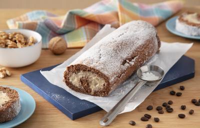 Foto da Receita de Rocambole de Chocolate com Café. Observa-se o rocambola sobre uma tábua azul escura e uma superfície de madeira. Grãos de café decoram a foto, assim como nozes