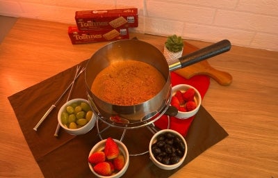 Fotografia em tons quentes com uma panela de fondue ao centro. Dentro da panelinha existe uma receita de fondue de cor caramelo feita com TOSTINES Especiarias. Ao lado, para acompanhar, existe um potinho com morangos e outro com uvas.