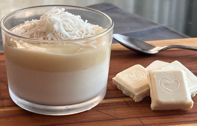 Imagem mostra um copo pequeno transparente com um creme branco por baixo e um creme bege por cima com coco ralado. Uma colher prateada está ao lado, junto de quadradinhos de chocolate branco. Tudo está apoiado em uma tábua de madeira.