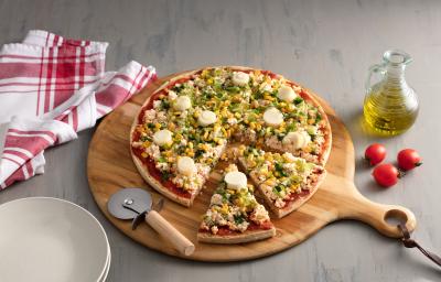 Fotografia em tons de vermelho em uma bancada de madeira cinza com uma tábua de pizza de madeira com a pizza em cima decorada com escarola e ricota. Ao lado, um vidro de azeite, tomates e um cortador de pizza.