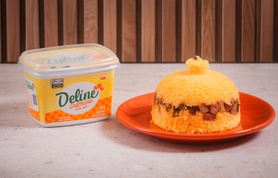Foto de um cuscuz recheado com charque, servido em um prato laranja, ao lado de uma embalagem de margarina deline