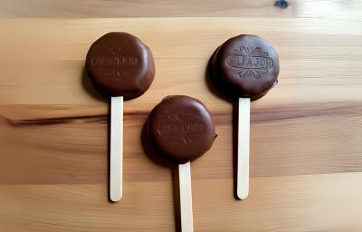 Fotografia em tons de madeira clara. Em cima da mesa existe 3 alfajores de chocolate meio amargo espetados em um palito de picole