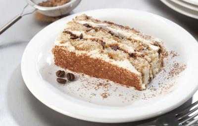 Fotografia em tons de branco, marrom e cinza, com prato branco ao centro com porção de bolo fatiado, no entorno garfo, peneirinha com chocolate em pó e pratos brancos empilhados, tudo sobre bancada em tons de cinza. Fotografia em tons de branco, marrom e cinza, com prato branco ao centro com porção de bolo fatiado, no entorno garfo, peneirinha com chocolate em pó e pratos brancos empilhados, tudo sobre bancada em tons de cinza.