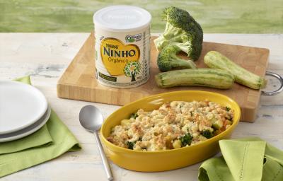 Fotografia em tons de verde e branco de uma bancada branca sobre ela um recipiente oval amarelo com o crumble de legumes. Ao lado um paninho verde, dois pratos brancos e uma colher. Ao fundo uma tábua de madeira com uma lata de NINHO, brócolis e abobrinha