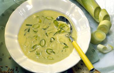 Fotografia de um prato branco fundo com um caldo de alho poró, com uma colher de cabo amarelo apoiada sobre a sopa, que tem pedaços de alho poró em cima. Ao redor, um alho poró inteiro, duas fatias de pão, uma taça vazia e grãos, sobre uma mesa verde. Fotografia de um prato branco fundo com um caldo de alho poró, com uma colher de cabo amarelo apoiada sobre a sopa, que tem pedaços de alho poró em cima. Ao redor, um alho poró inteiro, duas fatias de pão, uma taça vazia e grãos, sobre uma mesa verde.