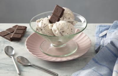 Foco da receita de MOÇA Sorvete de Flocos. Observa-se uma tacinha individual com três bolas do sorvete, servida com dois quadradinhos do chocolate Nestlé Classic meio amargo. A taça está sobre um prato de sobremesa rosinha.