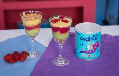 Fotografia em tons claros de duas taças de vidro com os cremes coloridos intercalados com o biscoito nesfit. As taças estão sobre dois panis, azul e roxo, ao lado tem a lata de ascenda e três morangos inteiros.