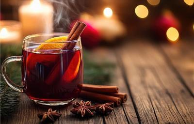 Fotografia de uma xícara de vidro com vinho quente, rodela de laranj e um pau de canela dentro. Ao lado tem três paus de canela e três cravos da índia sobre uma mesa de madeira.