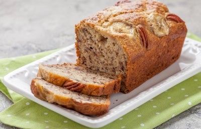 Fotografia em tons claros de um bolo de banana integral feito em forma de bolo inglês, com bananas e castanha decorando o bolo, que está sobre um apoio retangular branco, com duas fatias cortadas. Fotografia em tons claros de um bolo de banana integral feito em forma de bolo inglês, com bananas e castanha decorando o bolo, que está sobre um apoio retangular branco, com duas fatias cortadas.