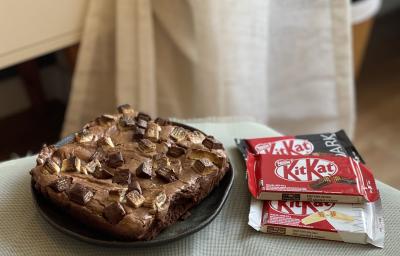Foto aproximada de um Brownie de KitKat, em formato quadrado, sobre um prato preto. Ao lado dele há três KitKtas, um ao leite, outro dark e outro white.