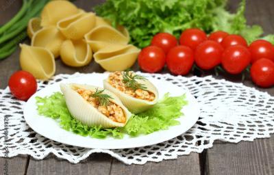Fotografia de duas unidades de macarrão tipo concha recheado com creme de palmito sobre uma folha de alface, que está em um prato redondo branco e pequeno. Ao fundo, algumas unidades de macarrão tipo concha crus e ao lado, tomates cereja. Fotografia de duas unidades de macarrão tipo concha recheado com creme de palmito sobre uma folha de alface, que está em um prato redondo branco e pequeno. Ao fundo, algumas unidades de macarrão tipo concha crus e ao lado, tomates cereja.