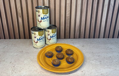 Seis brigadeiros caramelizados em forminhas azuis sobre prato amarelo, com três latas de LEITE MOÇA® ao fundo.