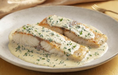 Filé de peixe branco dourado servido no molho cremoso de ervas, apresentado em prato fundo branco com acabamento sofisticado.