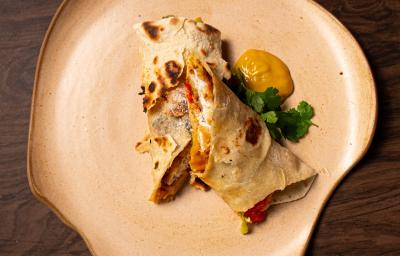 Fotografia de um wrap de moqueca cortado na metade, e cada metade sobre a outra. Ao lado do wrap tem um molho de mostarda e salsinha.