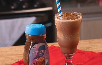 Fotografia de um copo de vidro grande com frozen de café, ao lado de uma embalagem de nescafé gelado, sobre um pano vermelho e uma mesa de madeira. Dentro do copo tem um canudo azul e branco apoiado. Fotografia de um copo de vidro grande com frozen de café, ao lado de uma embalagem de nescafé gelado, sobre um pano vermelho e uma mesa de madeira. Dentro do copo tem um canudo azul e branco apoiado.