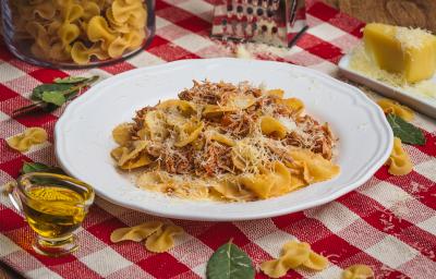 Foto de uma mesa com uma toalha xadrez vermelha e branco. Sobre ela já macarrões crus, um pedaço de queijo, um ralador, um pote com azeite e algumas folhas. No centro há um prato branco com a receita de Farfalle com Ragú de Frango.