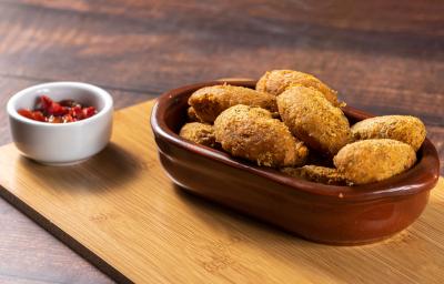 Fotografia de uma travessa marrom com vários bolinhos fritos em tons dourados por cima de uma tábua de madeira. Ao lado, um pequeno recipiente branco com um molho de pimentas vermelho.