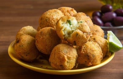 Em cima de uma mesa, prato amarelo com porção de bolinhos de bacalhau dourados e crocantes. No lado direito, há um bolinho aberto ao meio com bacalhau cremoso. Em cima de uma mesa, prato amarelo com porção de bolinhos de bacalhau dourados e crocantes. No lado direito, há um bolinho aberto ao meio com bacalhau cremoso.